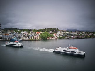 Bilde av to hurtobåter i forgrunnen av bildet. Bildet viser og husstander og annen bebyggelse langs kysten. Himmelen er grå