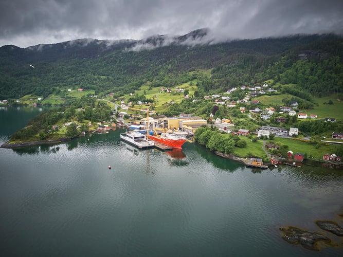 Fjord og fjell, skyer dekker toippen av fjellene i bakgrunnen. Tettbygd område med Fjellstrand verft i forgrunnen. Industribygg og skip som ligger til kai. Boligområder langs vei og oppe i lien.