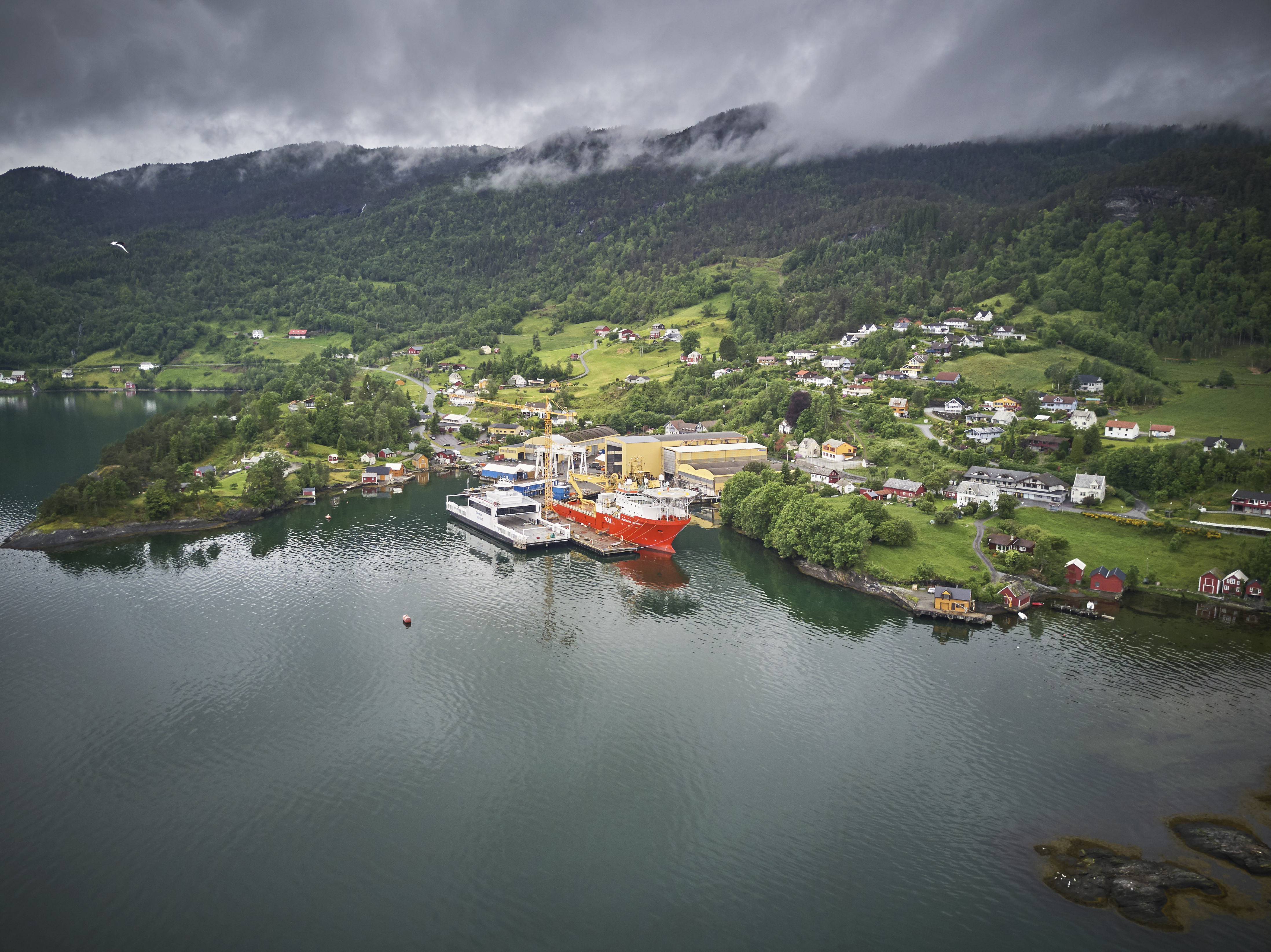Fjord og fjell, skyer dekker toippen av fjellene i bakgrunnen. Tettbygd område med Fjellstrand verft i forgrunnen. Industribygg og skip som ligger til kai. Boligområder langs vei og oppe i lien.